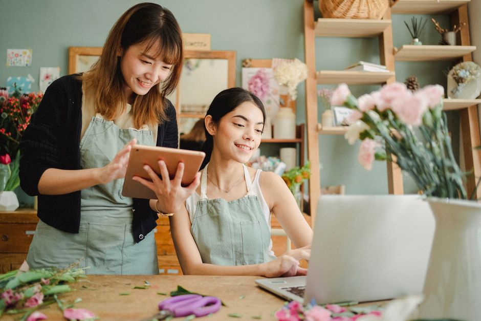 Two women working in a florist shop, one standing and smiling while holding a tablet, and the other seated at a wooden work table with a laptop, surrounded by fresh flowers and floral supplies. The se