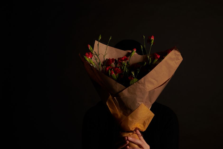 A person holding a bouquet of fresh red and pink flowers wrapped in brown kraft paper and beige tissue paper, with a black ribbon tying the bouquet. The person's face is obscured by the flowers, and t