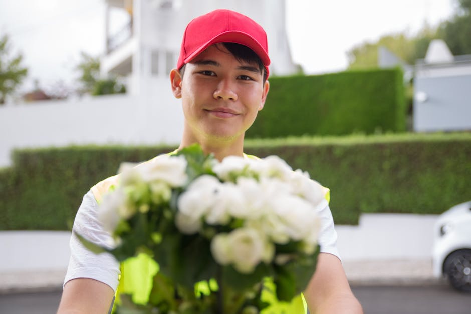 A young delivery person wearing a bright red cap and a high-visibility yellow vest is standing outdoors on a street in Greenford, holding a fresh floral gift consisting of white roses and green foliag