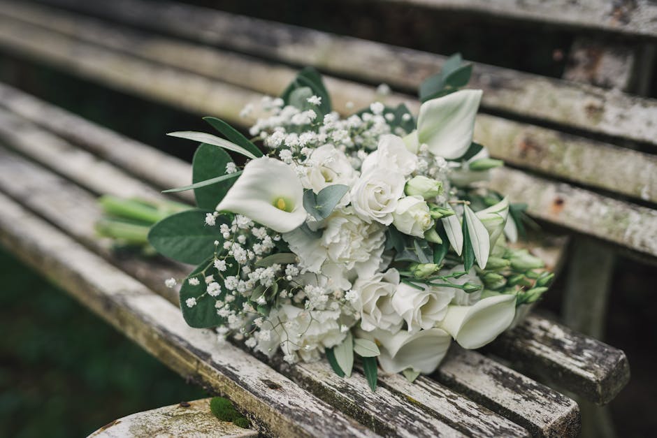 A bouquet of fresh white flowers, including calla lilies, roses, and eustoma, arranged with eucalyptus leaves, baby's breath, and other greenery. The arrangement is wrapped in white paper with a soft 