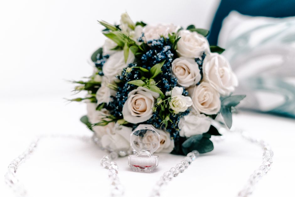 A bridal bouquet featuring creamy white roses, accented with small green buds and lush green foliage, arranged in a rounded, hand-held style. The bouquet rests on a white surface, with soft lighting e