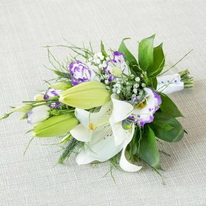An elongated, hand-tied bouquet lies at a diagonal across a softly textured, light beige fabric, the neutral background echoing the calm skies over Greenford on a clear morning. The composition is led by several large white Oriental lilies, fully opened so that their broad, gently recurved petals form star-shaped blooms. Each lily reveals a cluster of yellow-orange stamens dusted with pollen, surrounded by faint freckles and subtle shading at the throat, suggesting both delicacy and strength. Between them, sleek, chartreuse-green lily buds stand upright, tightly closed and smooth, hinting at blossoms still to come. Weaving through this structure is a profusion of Lisianthus in white and purple: frilled, cup-shaped flowers whose creamy bases transition into vivid violet edges, some deep and dramatic, others softened into lavender tones that lighten the overall effect. Tiny gypsophila, or baby's breath, appears in fine, branching sprays, scattered like a delicate mist, filling the spaces between larger blooms and adding a breathable, airy softness to the design. The bouquet is cradled by an abundance of greenery-broad, glossy dark green leaves give weight and contrast, while finer, feathery foliage and slender, slightly spiky blades add a touch of wild movement, reminiscent of the natural growth along nearby Brent Valley Park. At the base, the stems are gathered cleanly and wrapped in smooth white satin ribbon, accented with small, pearl-like beads that catch the soft, diffused light, suggesting this is a bouquet intended for a bride, a bridesmaid, or an important milestone celebration. The overall mood is elegant yet fresh, as though it has just been prepared for same-day delivery across Greenford, the kind of refined arrangement a local florist would recommend for a wedding, anniversary, or sophisticated thank-you gift.