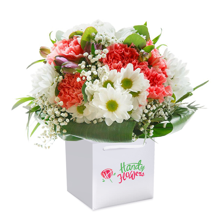 Expertly styled by a Greenford florist, this vibrant bouquet is presented upright in a spotless square white gift bag, illuminated against a clear white background that calls attention to every petal and leaf. The design balances soft coral and lively peach carnations-renowned for their plush, ruffled texture-and striking white daisy-like blooms whose golden-green centers provide a playful, cheery contrast. Sprigs of delicate, cloud-like baby's breath dart among the fuller flowers, weaving airy movement into the composition. A rich collar of deep, glossy green leaves forms the bouquet's base, reminiscent of the abundant greens found in the gardens around Greenford Broadway. Along the edge, a few unopened buds brushed with hints of purple and pink lend depth and anticipation, as though capturing a fleeting moment in spring. The bouquet's rounded silhouette is meticulously achieved, projecting abundance and elegance while hinting at the care and precision behind every stem. With sturdy white ribbon handles for easy carrying and tasteful local branding, the presentation is perfect for gifting. This image radiates freshness, refined artistry, and the heartfelt tradition of flower-giving-a true representation of the quality and convenience of flower delivery available throughout Greenford.