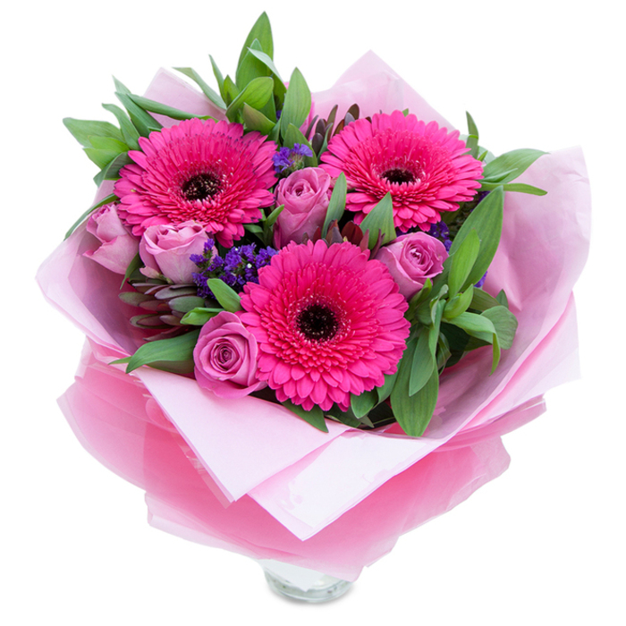 I see this bouquet as an intimate celebration of pinks and purples, composed with an eye for both drama and tenderness. Dominant fuchsia Gerbera daisies open wide at the heart of the design, their vivid petals forming sunburst shapes around deep, dark centers that lend a dramatic focal point. Interwoven are several roses in a gentle lavender-pink or dusty rose, their petals still snug in fresh buds that promise a slow, elegant unfurling. Small clusters of purple statice or limonium peek through the spaces, bringing a peppering of cool violet and delicate texture that feels almost meadow-like. The green foundation-broad, glossy foliage reminiscent of salal and the grey-green sweep of eucalyptus-frames the flowers with a crisp, verdant profile and a faint, dewy aroma. The bouquet is wrapped thoughtfully: an inner layer of soft matte pale pink tissue cushions the stems, while an outer wrap of translucent, slightly crinkled fuchsia-pink cellophane imparts a subtle shimmer and a unified colour story; the folds are neat, the presentation considered. A rim of clear glass at the bottom hints that this bouquet is arranged for immediate display, perfect to sit on a kitchen island after a quick stop at Greenford Broadway or to arrive for a special moment near Ealing Broadway. Bathed in soft indoor light, the composition reads as optimistic and caring-the kind of arrangement a local florist would choose to send with birthday wishes, a thank-you, or gentle congratulations to someone you want to make feel cherished.