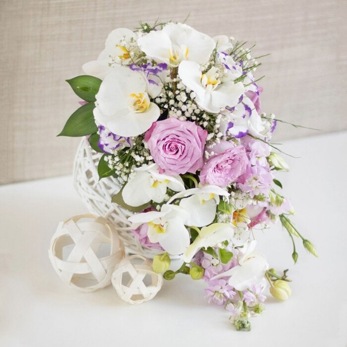 An elegant, sculptural floral arrangement is captured against a softly textured neutral wall, as if styled for a refined Greenford wedding or anniversary dinner near Perivale Park. The arrangement is built within a distinctive white spherical vase, created from interlaced strands that form an open lattice, like an oversized, artistically woven ball of yarn. This airy base lifts the blooms and adds a contemporary yet gentle character to the scene. From it, long, arching stems of pristine white Phalaenopsis orchids extend, their wide petals smooth and pristine, each flower centred with detailed touches of yellow and soft amber that bring a subtle warmth to the design. Surrounding the orchids are full, ruffled roses in delicate blush pink and lavender shades, many at the peak of bloom, their petals unfurling in rich layers that suggest softness and fragrance. Intertwined through the larger blooms, clouds of white baby's breath add a fine, almost cloud-like texture, lightening the silhouette and drawing the eye throughout the arrangement. Smaller, frilled blossoms-likely lisianthus or eustoma-appear in white with faint purple edges, lending an extra dimension of colour and detail. From the lower edges, stems of pale lilac trailing flowers spill gently downward, giving the arrangement a sense of relaxed movement, as though it might slowly cascade across a wedding table along Oldfield Lane. Glossy green leaves and finer, fern-like foliage emerge between the blossoms, grounding the pastels and providing fresh contrast. On the smooth white surface beside the vase, two decorative orbs in a soft cream tone echo the same open, woven pattern, one larger and one smaller, balancing the visual weight of the main piece. The soft, even lighting enhances every petal and strand, creating a tranquil yet festive atmosphere perfectly suited to weddings and special celebrations throughout Greenford.
