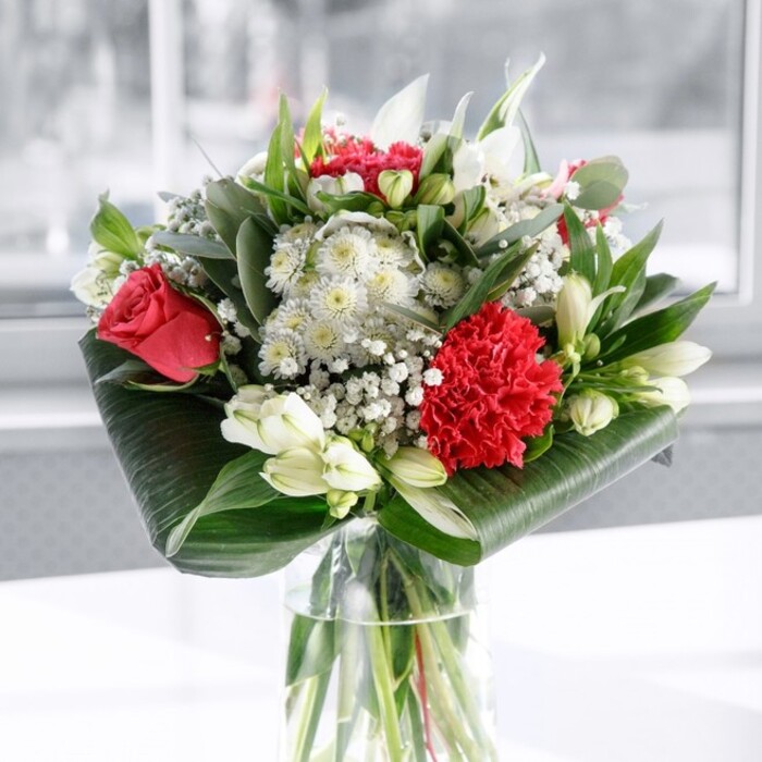 This image presents a thoughtfully crafted red and white bouquet arranged in a tall, clear cylindrical glass vase, the stems visible through the crystal-like water in an orderly cluster of fresh green. At the core of the design, velvety deep red roses unfurl with classic grace, each petal layered and smooth, suggesting enduring romance, while a large, intricately ruffled crimson carnation is positioned near the front as a textured centerpiece. Around these intense red tones, a harmony of white and cream blossoms creates balance: white Alstroemeria with slender petals and subtle inner speckles, full-headed white chrysanthemums, and several firm white lily buds that have yet to open, their smooth, closed forms hinting at slowly emerging fragrance. Between the larger flowers, delicate clouds of white gypsophila, or baby's breath, weave through the arrangement, their tiny blooms creating a gentle, airy veil that lightens the overall composition. Supporting this floral display is a rich frame of greenery-broad, glossy dark green leaves are expertly curled and wrapped around the base, forming a polished collar, while finer eucalyptus-like sprigs and pointed foliage interlace between blossoms, offering varied shapes and tones of green reminiscent of the leafy paths around Brent Lodge Park. The bouquet rests on a clean white surface in front of a softly blurred, bright backdrop that suggests natural daylight streaming through a window in a Greenford home. The lighting is even and calm, enhancing the saturation of the reds and the calm purity of the whites, without overshadowing the subtle textures. The overall feeling is one of poised celebration and heartfelt sentiment, ideal as a centerpiece for a family gathering, a romantic gesture delivered across Greenford, or a refined gift to bring warmth and elegance to an office reception desk.