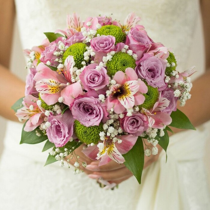 A breathtaking, close-up bridal bouquet is gently held against the white wedding dress of a newlywed in Greenford, highlighting the careful artistry of local florists. The main feature is a gorgeous gathering of lavender and blush pink roses, some of which still carry fresh dew, their petals bursting with springtime promise. Round, zesty green button chrysanthemums add a modern twist, their unique texture contrasting beautifully against the traditional roses. Elegant alstroemeria lilies with soft pink petals streaked in dark pink and yellow drift through the bouquet, imparting a subtle exotic charm. Abundant clusters of delicate white baby's breath weave a halo of softness and ethereal light around the bolder blooms, creating a sense of weightlessness. Deep green leaves curve around the bouquet's base, setting off the colours and lending a touch of local garden vibrancy. The focus on the bride's hands and her simple ring evokes personal commitment and gentle anticipation, all lovingly set against the textured garment. This bouquet perfectly captures Greenford's blend of contemporary taste and classic wedding tradition, a fresh and radiant accessory for spring or summer weddings and an inspiring option for event décor in the Greenford area.