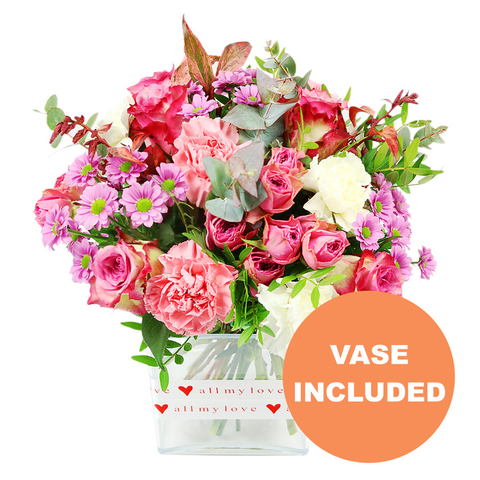 Centred in the frame and set against a clean white background, a vibrant mixed bouquet rises from a clear, square glass vase, its rich colours and interplay of textures immediately drawing the eye. The dominant blooms are pink roses, some in striking shades of deep fuchsia and magenta, others in gentler blush and pastel tones, each rose captured at a slightly different stage of opening so that the bouquet feels alive and evolving. Around them, generous carnations in tones of coral, peach-pink, and creamy white unfurl their signature ruffled petals, providing a plush, layered texture that gives the arrangement a sense of luxurious volume. Punctuating the pinks and whites are daisy-like chrysanthemums in lavender and soft purple, their bright lime-green centres catching the light and adding an unexpectedly modern twist to the classic romantic palette. A well-balanced selection of greenery supports the flowers: rounded, silvery-green eucalyptus leaves, darker green spray foliage, and the occasional reddish-brown leaf that suggests the warm, earthy hues you might notice in Greenford's gardens as the seasons turn. The stems are neatly gathered in the vase, visible through the clear glass and resting in fresh water, reflecting the kind of careful conditioning that local florists rely on for long-lasting arrangements. Around the lower part of the vase, a white band is printed with small repeating red hearts and the phrase all my love in flowing red script, making the bouquet feel distinctly suited to romantic moments, Mother's Day surprises, or thoughtful thank-you gestures for neighbours and friends around Greenford. In the lower right corner of the image, an orange circular overlay carries the words VASE INCLUDED in bold white capitals, highlighting that this is a complete, ready-to-display floral gift, thoughtfully composed in the style of Greenford Flowers for easy home or office placement.