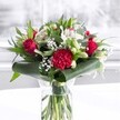 A lush, romantic bouquet stands proudly in a clear cylindrical glass vase, its waterline revealing a neat cluster of freshly trimmed green stems that speak of same-day freshness in Greenford. At the heart of the design, velvety deep red roses unfurl in perfect spirals, their petals rich and layered like classic love letters, while a ruffled crimson carnation nestles at the front, its densely frilled petals adding a sense of drama and tactile depth. Around these passionate reds, soft white blooms create a gentle halo: alabaster Alstroemeria with slender, speckled petals, creamy white chrysanthemums, and a few tightly closed white lily buds that hint at perfumed blossoms still to come. Airy clouds of white gypsophila, or baby's breath, appear like tiny stars sprinkled throughout, giving the bouquet a light, almost floating quality, as if a cool breeze from nearby Ravenor Park had passed through. Lush greenery frames the flowers with intention and care: broad, glossy dark green tropical leaves are artfully curled and wrapped around the base, forming a collar that grounds the arrangement, while slimmer eucalyptus-style foliage and pointed leaves weave upwards, adding layered shades of green and a subtle woodland texture. The vase rests on a clean white surface, perhaps a kitchen table in a Greenford home near the Broadway, bathed in soft, even daylight that brightens every petal and leaf. The overall mood is warm and celebratory yet refined, ideal as a heartfelt anniversary surprise, a Mother's Day gift, or a thoughtful centerpiece for a family gathering, conveying affection, gratitude, and a sense of welcome with every bloom.