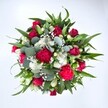 Viewed from directly above against a clean white background, this generous bouquet forms a lush, almost perfectly round circle of colour, as if a little garden has been gathered into a single, overflowing armful. At the centre, deep crimson red roses anchor the design, some still held in tight buds, others beginning to unfurl their velvety petals, suggesting romance, promise, and quiet elegance. Around them, ruffled carnations in rich fuchsia and ruby red spill out in soft waves, each frill catching the light and adding a playful, festive energy. Between these bold blooms, clusters of small white spray chrysanthemums - daisy-like and cheerful - light up the arrangement with their fresh, bright faces, like tiny stars scattered across a night sky. Threaded delicately through the bouquet are countless miniature white gypsophila blossoms, or baby's breath, creating a sheer, cloud-like veil that softens every edge. Supporting and shaping the flowers is an abundance of greenery: broad, pointed dark green leaves, likely from lilies or alstroemeria, reach outward to give structure and drama, while rounded, silvery-green eucalyptus leaves weave in and out, adding a contemporary twist and a suggestion of cool, herbal fragrance. The natural daylight in the photograph keeps every colour crisp and true, from the rich reds to the clear whites and muted greens, so the whole bouquet appears freshly arranged on a workbench in Greenford, ready to be delivered across the local area for a Valentine's surprise, an anniversary, or simply to lift someone's spirits with a vivid expression of affection and vitality.
