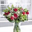 A richly detailed photograph shows a classic mixed bouquet of red roses, red carnations, white alstroemeria, and clouds of dainty baby's breath, gathered tightly in a clear glass vase set on a bright white tabletop. The deep crimson roses are the stars, some fully open with velvety, spiralling petals, others still in smooth, promising buds, suggesting romance that will unfold over days. Around them, ruffled red carnations add plush texture and generous volume, each frilled petal catching the soft daylight. Tucked between the bold reds are clusters of white alstroemeria, many still in bud with pale green-tipped petals, while a few blossoms have opened to reveal their trumpet-like shape and faint striping, bringing a gentle, elegant contrast. Fine sprays of gypsophila, or baby's breath, thread through the arrangement, like tiny white pearls or a soft mist hovering among the larger blooms, lending a dreamy, cloudlike softness. The flowers are cradled by a collar of lush greenery: broad, glossy dark green leaves frame the base, while slimmer, lighter green stems and leaves weave upward, adding lift and movement to the overall silhouette. Through the sides of the cylindrical glass vase, the neatly cut stems are clearly visible, fanning into clear water that reinforces the sense of freshness, as if the bouquet has just been arranged by a local Greenford florist. The setting is airy and modern, with a large window in the softly blurred background diffusing pale natural light across the scene. Outside the window, the view is rendered in muted, almost grayscale tones-perhaps reminiscent of a quiet Greenford side street near Ravenor Park-so that the vibrant reds, whites, and greens of the bouquet stand out boldly. The atmosphere feels warmly celebratory and timeless, ideal for marking an anniversary, Valentine's gesture, or a heartfelt thank-you delivery anywhere across Greenford and the surrounding neighbourhoods.